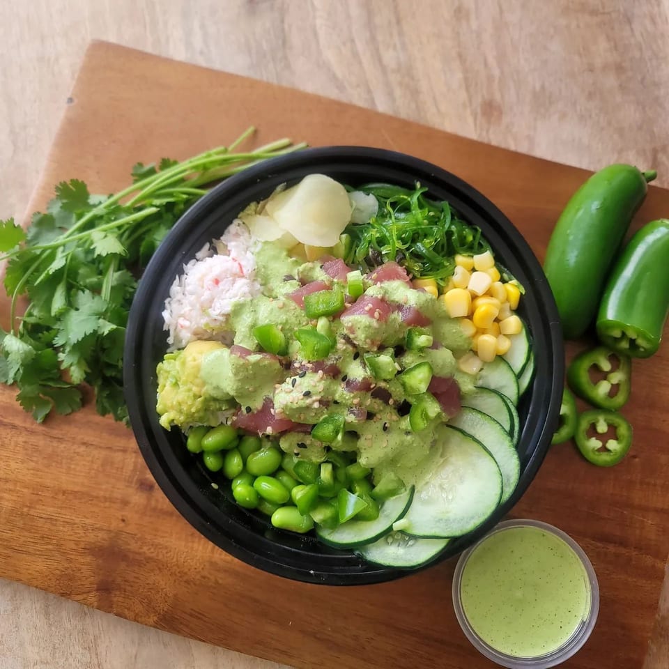Oh Poke bowl with tuna, avocado, edamame, cucumber, corn, and green sauce on a wooden board.