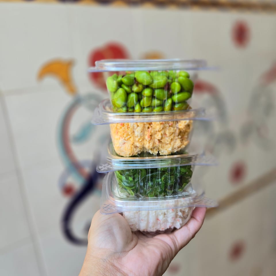 Hand holding a stack of four clear containers with edamame, crab salad, seaweed, and another poke ingredient.