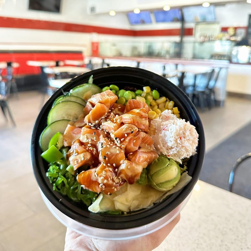 A hand holds a poke bowl with salmon, crab, avocado, cucumber, edamame, and corn.