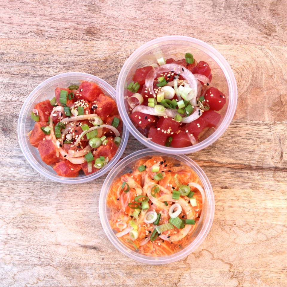 Three plastic containers of Oh Poke, two with tuna and one with salmon, on a wooden surface.